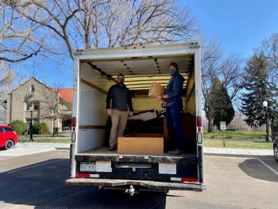 Two men stand inside the Partners in Housing box truck with masks on, posing with wooden shelving. This is a delivery of furniture from volunteers to a family in the Self Sufficiency Program.