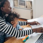 Woman working on paperwork with a laptop while holding her child on her lap.