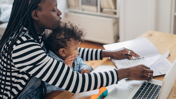 Woman working on paperwork with a laptop while holding her child on her lap.