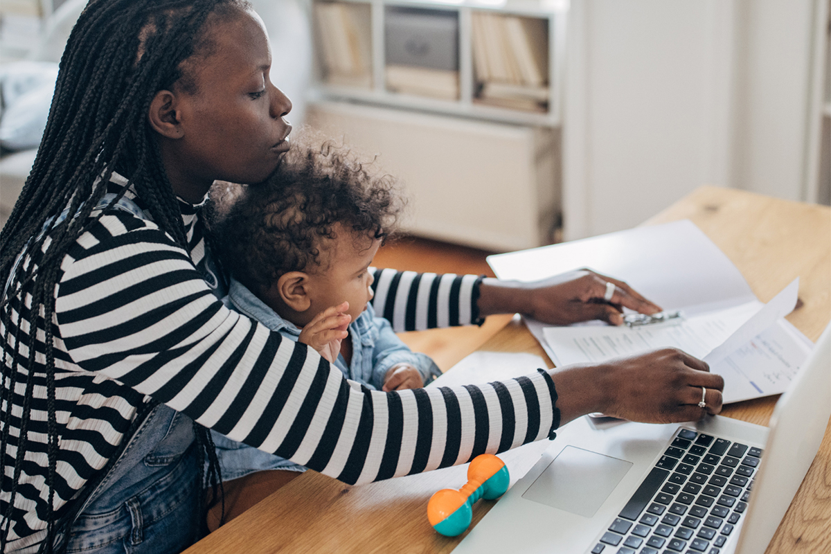 Woman working on paperwork with a laptop while holding her child on her lap.